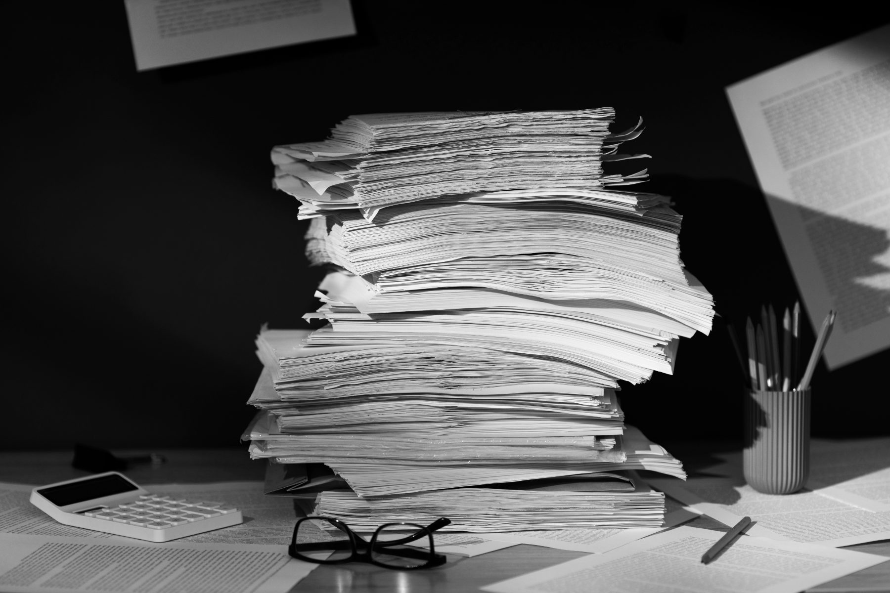 A tall stack of papers on a desk beside a calculator and a pencil cup, in black and white