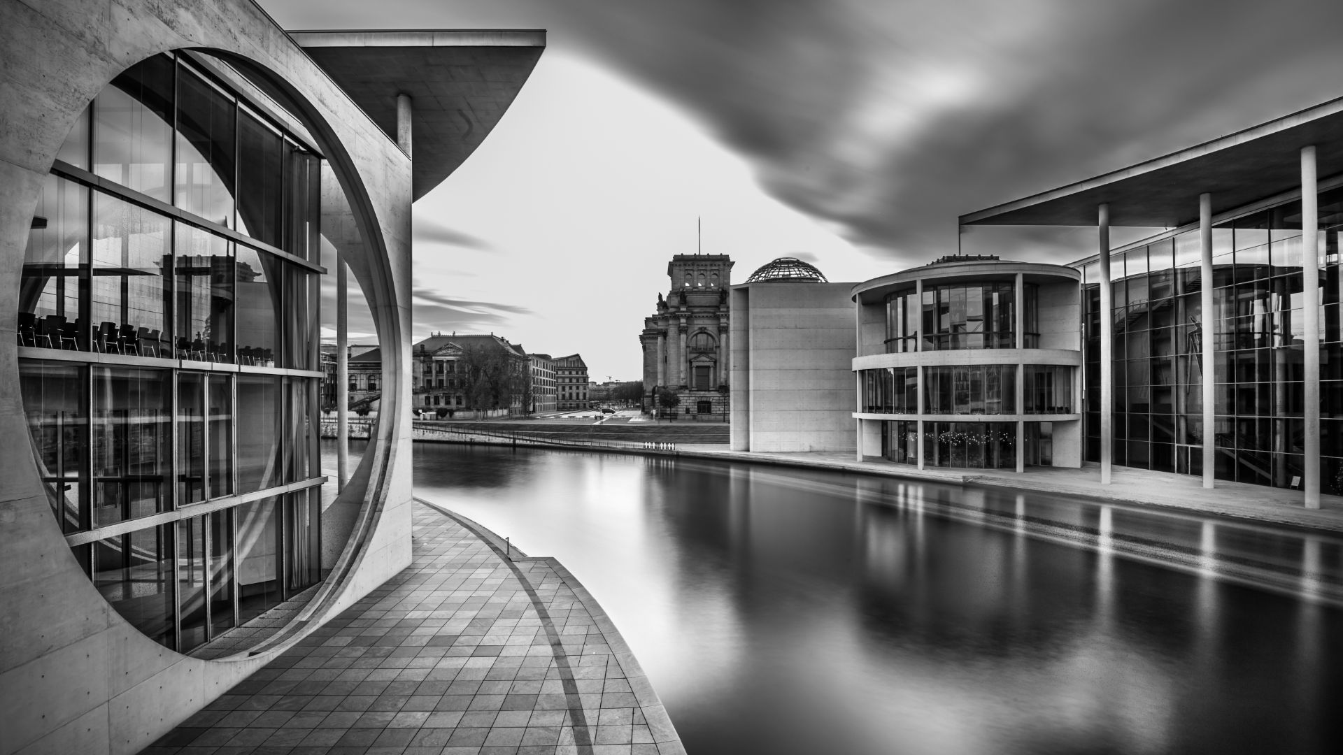 Modern government buildings along a still river under a long-exposure sky, in black and white