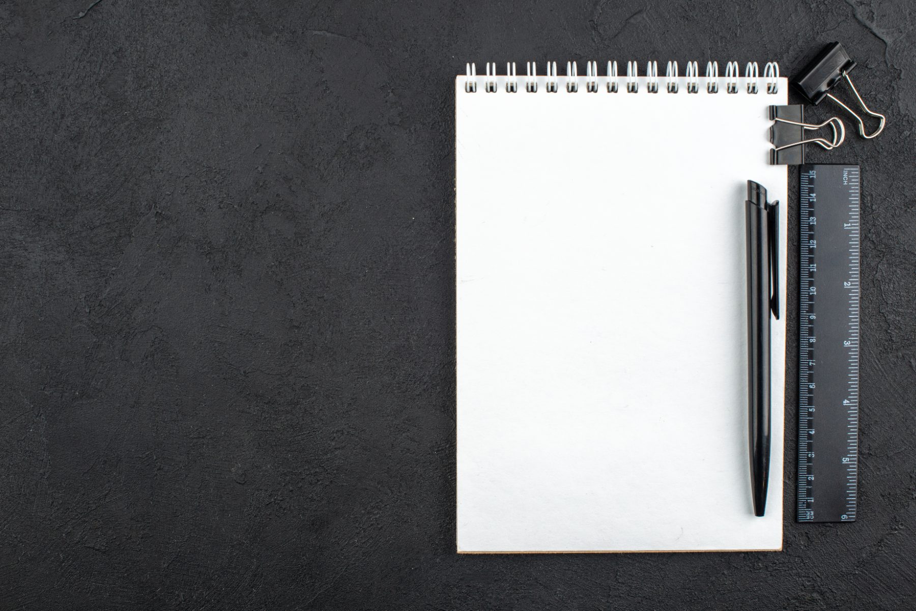 A pen, notepad, ruler and binder clips on a dark surface
